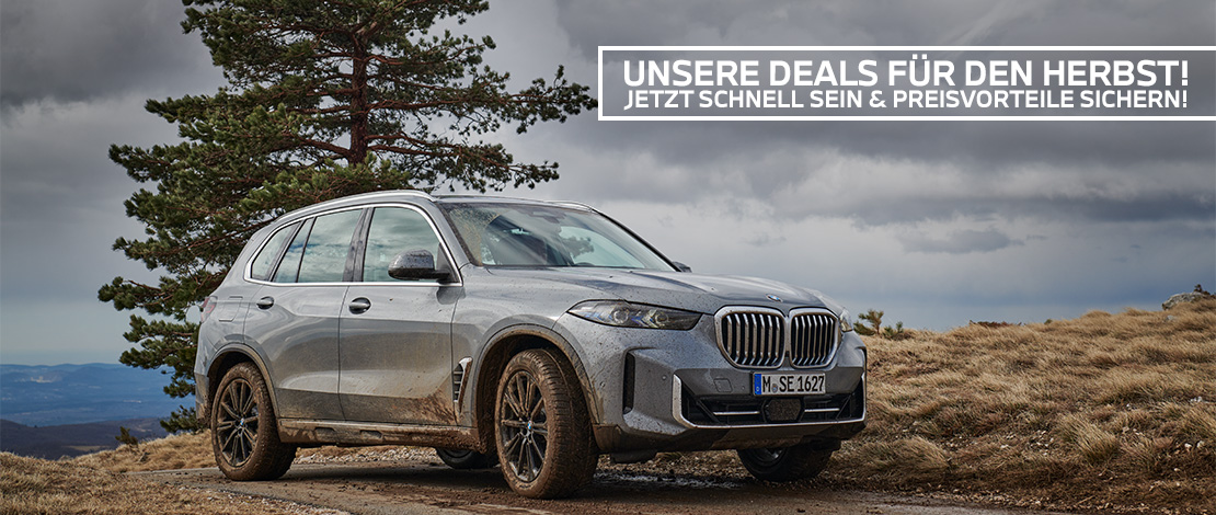 Das Bild zeigt ein graues BMW SUV (wahrscheinlich ein X5), das leicht mit Schmutz bedeckt ist. Das Auto steht auf einem Schotterweg in einer natürlichen, herbstlichen oder bergigen Umgebung. Im Hintergrund sind eine große Kiefer und eine weite Landschaft unter einem bedeckten, dramatischen Himmel zu sehen. Text im Bild: Über dem Auto befindet sich ein weißes Banner mit schwarzer Schrift. Der Text lautet: "UNSERE DEALS FÜR DEN HERBST! JETZT SCHNELL SEIN & PREISVORTEILE SICHERN!"
