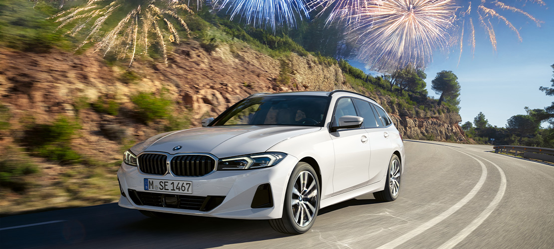 Ein weißer BMW 3er Touring fährt dynamisch eine kurvenreiche Straße entlang, die von bewaldeten Hügeln gesäumt ist. Das Bild ist als Werbegrafik gestaltet: In der oberen linken Ecke prangt ein großes, blaues Prozentzeichen (%), während in der oberen rechten Ecke ein Feuerwerk vor blauem Himmel zu sehen ist. Ein schmaler weißer Rahmen fasst das gesamte Motiv ein und signalisiert besondere Rabattaktionen im Rahmen der Jahresstart-Kampagne.