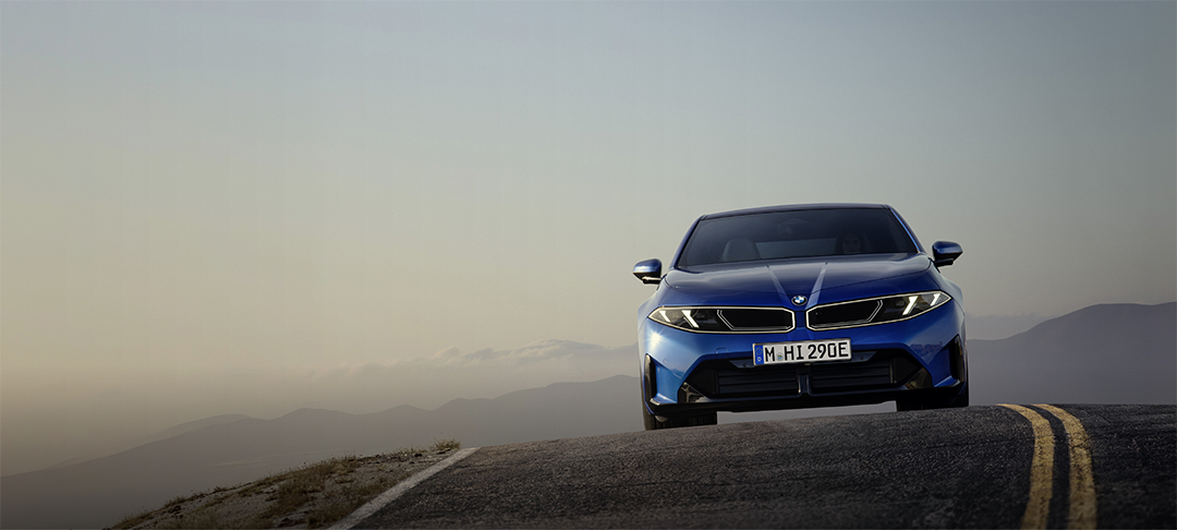 Neuer vollelektrischer BMW i3 in leuchtend blauer Lackierung aus der Frontperspektive auf einer Bergstraße. Die markante BMW Niere, die schmalen LED-Scheinwerfer und die sportlich gestaltete Frontpartie stehen im Mittelpunkt. Im Hintergrund erstreckt sich eine weitläufige Berglandschaft im sanften Licht der Abenddämmerung