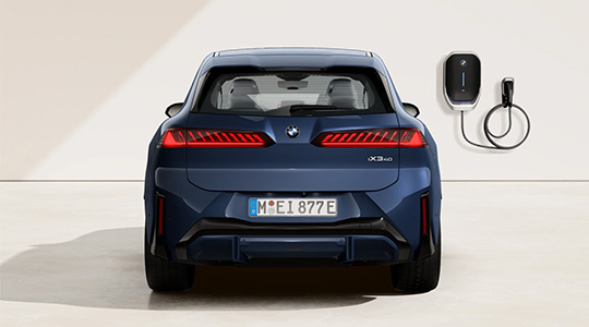Heckansicht des dunkelblauen BMW iX3 40 (Modellgeneration Neue Klasse) im Studio mit leuchtenden, markanten LED-Heckleuchten. Rechts an der weißen Wand ist eine BMW Wallbox mit aufgewickeltem Ladekabel montiert.
