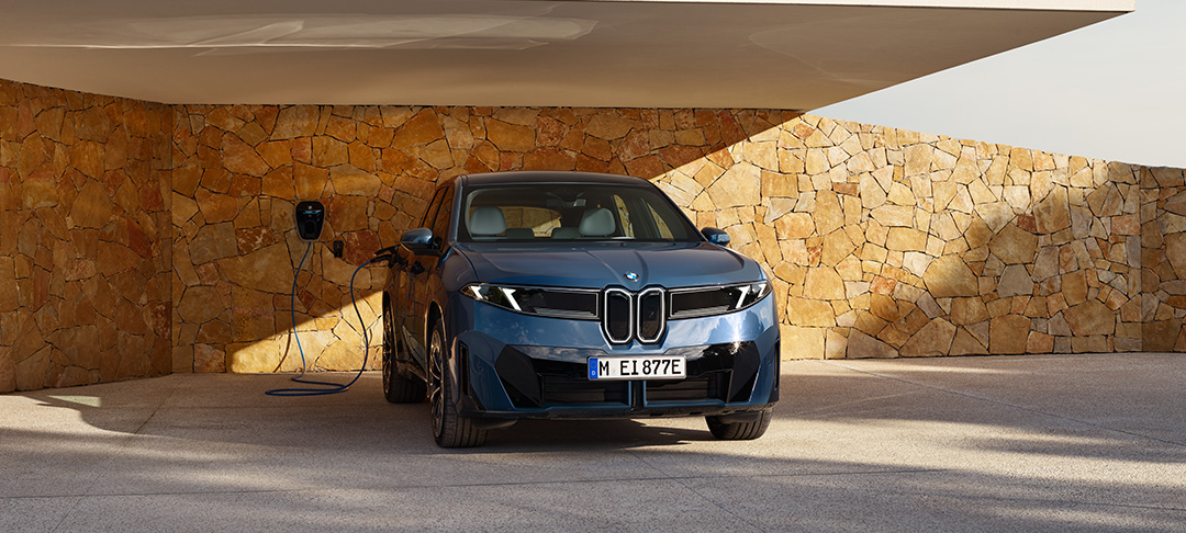Ein blauer BMW Elektro-SUV der „Neue Klasse“ Konzeptreihe parkt unter einem modernen Carport und ist über ein Ladekabel an einer Wallbox an einer Natursteinwand angeschlossen.