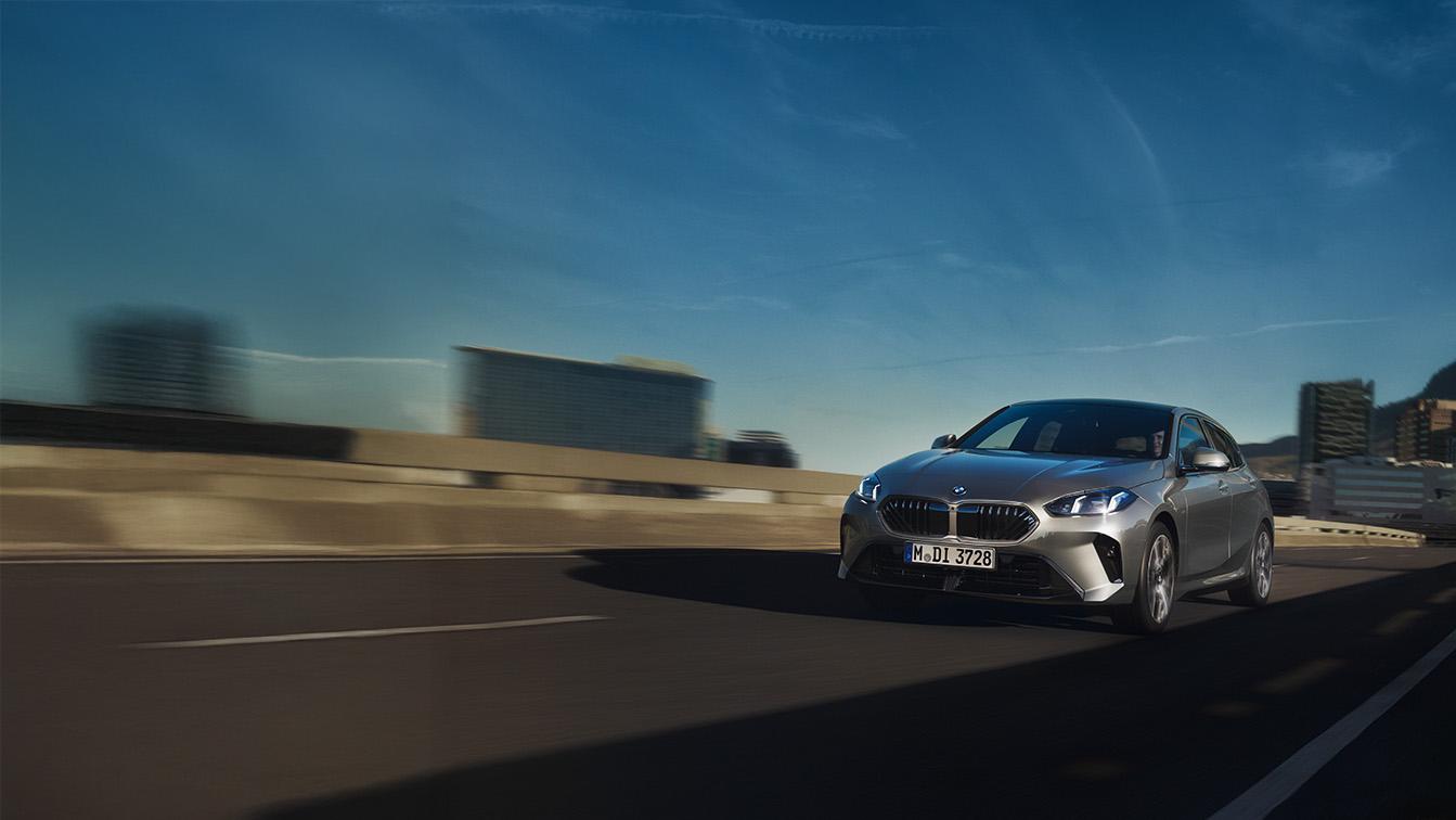 Ein silberner BMW 120 fährt bei strahlendem Sonnenschein unter einem tiefblauen Himmel auf einer Schnellstraße direkt auf den Betrachter zu. Während das Fahrzeug in seinem metallischen Glanz gestochen scharf abgebildet ist, verschwimmen der graue Asphalt und die städtische Silhouette im Hintergrund in starker Bewegungsunschärfe, was ein intensives Gefühl von Geschwindigkeit vermittelt. Die markante Front mit den hell leuchtenden LED-Scheinwerfern und dem typischen Doppelkühlergrill steht dabei im Fokus, während harte Schatten auf der Fahrbahn die kraftvolle Dynamik der Szene unterstreichen.