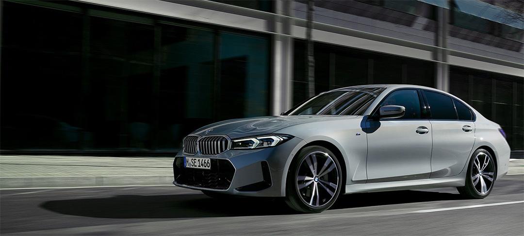 BMW 318i Limousine Angebot Ein silberner BMW 3er Sedan fährt auf einer Straße, vermutlich in einer Stadt, da im Hintergrund moderne Gebäude mit großen Fenstern zu sehen sind. Das Auto ist von vorne rechts leicht schräg aufgenommen und bewegt sich scheinbar nach links im Bild. Die Tagfahrlichter sind eingeschaltet und spiegeln sich leicht im Asphalt. Die Felgen sind schwarz-silber. Das Kennzeichen lautet 'M SE 1466