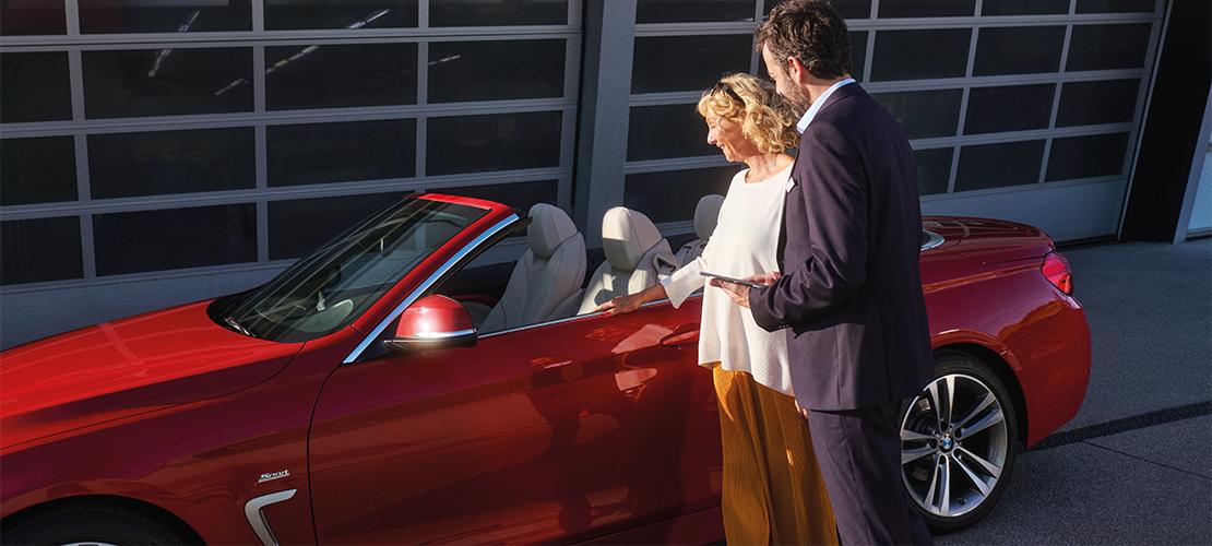 Eine lächelnde Frau steht neben einem roten BMW Cabriolet und unterhält sich mit einem Mann, der ein Tablet hält. Im Hintergrund ist eine Garagenwand zu sehen