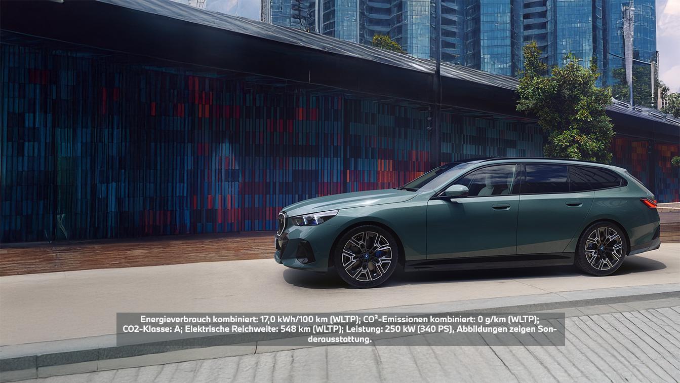 Ein dunkelgrüner BMW i5 Touring Kombi ist seitlich, leicht schräg von vorne gesehen, auf einem gepflasterten Weg vor einem modernen Gebäude mit farbiger Fassade und gläsernen Hochhäusern abgebildet. Das Fahrzeug hat schwarze Felgen mit blauen Akzenten und getönte Scheiben. Ein Textoverlay informiert über die technischen Daten: Der kombinierte Energieverbrauch (WLTP) beträgt 17,0 kWh/100 km, die CO₂-Emissionen (WLTP) sind 0 g/km (CO₂-Klasse A), die elektrische Reichweite (WLTP) liegt bei 548 km, und die Leistung beträgt 250 kW (340 PS). Die Abbildungen zeigen Sonderausstattung.