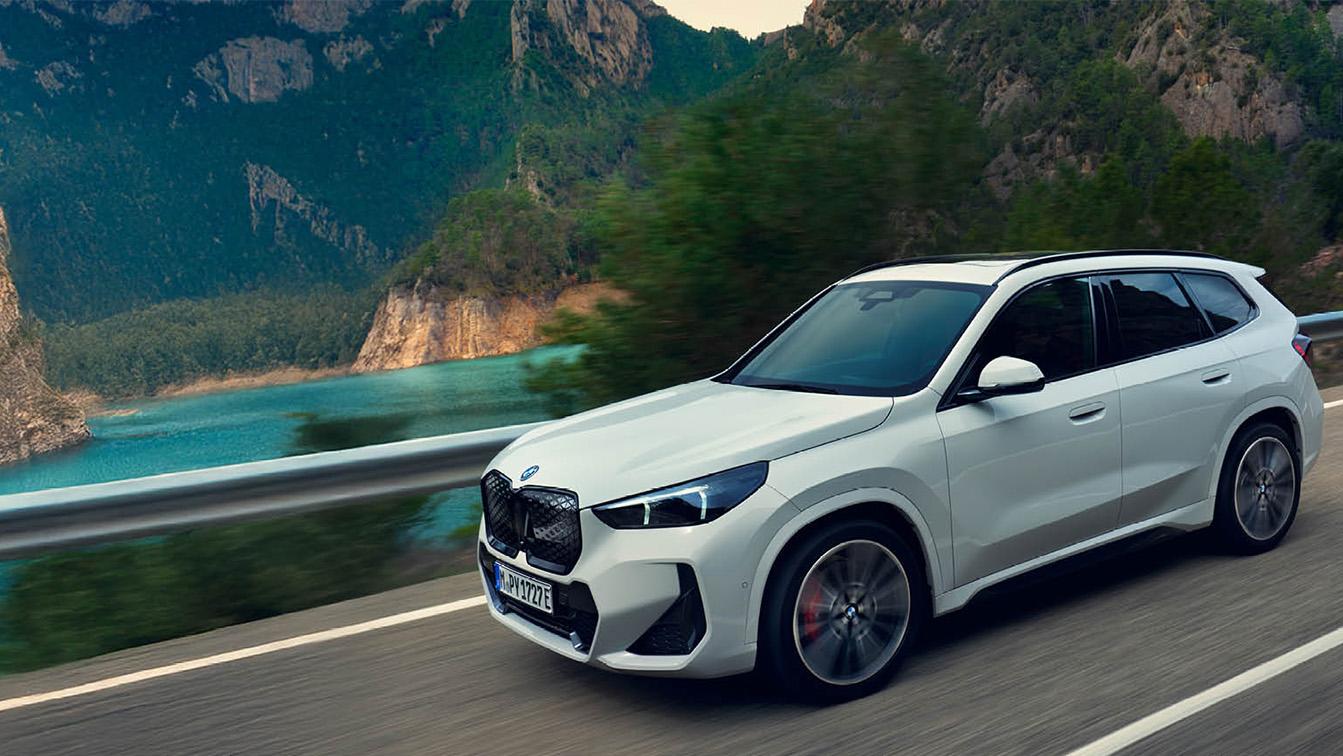 Ein weißer BMW iX1 fährt dynamisch über eine kurvige Bergstraße, die sich entlang eines tiefblauen Bergsees durch eine imposante Wald- und Felskulisse schlängelt. Das Elektro-SUV ist im Dreiviertelprofil von vorne zu sehen, wobei die leichte Unschärfe der Räder und der vorbeiziehenden Leitplanke die Geschwindigkeit und Leichtigkeit der Fahrt unterstreichen. Die Szenerie ist in ein sanftes, natürliches Tageslicht getaucht, das die moderne Linienführung des Wagens betont und eine Atmosphäre von Freiheit, technischem Fortschritt und Aufbruch in die Natur vermittelt.
