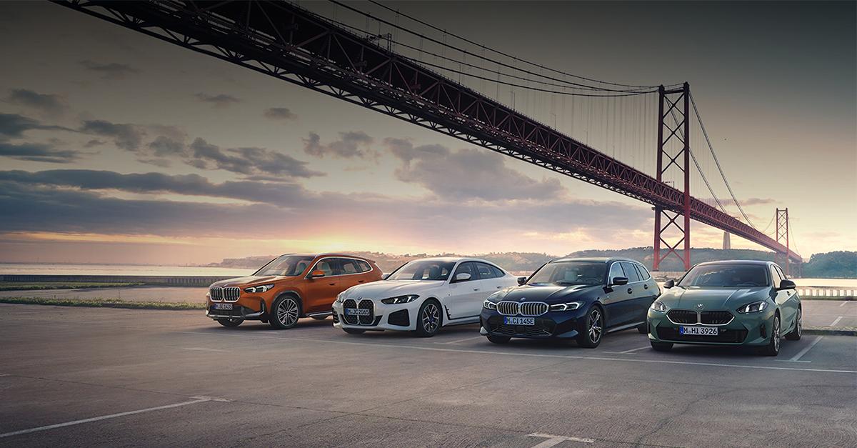 Vor der monumentalen Kulisse einer roten Hängebrücke präsentiert BMW vier aktuelle Modelle in der warmen Abendsonne. Auf einem weiten Parkplatz reihen sich ein orangefarbener X1, ein weißes 4er Gran Coupé, ein dunkelblauer 3er Touring und ein graugrüner 1er nebeneinander auf.  Die Szenerie am Wasser, die diagonale Stahlkonstruktion der Brücke und das goldene Licht verleihen der Aufnahme eine hochwertige, urbane Atmosphäre, die Vielseitigkeit und modernen Lifestyle ausstrahlt.