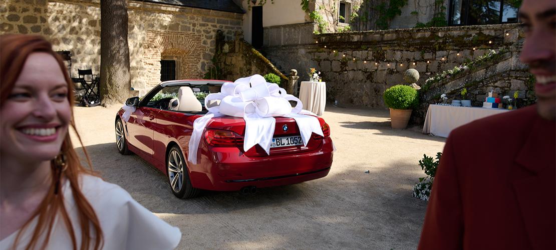 Ein rotes BMW Cabriolet ist mit einer großen weißen Schleife als Geschenk dekoriert und steht auf einem gepflasterten Hof vor einem historischen Gebäude. Unscharf im Vordergrund sind eine lächelnde Frau und ein Mann zu sehen.