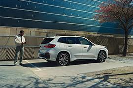 Der weiße BMW X1 ist von der Seite-hinten auf einem Parkplatz abgebildet. Ein Mann steht neben dem Auto und blickt auf sein Mobiltelefon. Die Umgebung zeigt eine moderne Gebäudefassade (hellblau und strukturiert) und einen Baum mit einigen rosa Blüten im Frühling