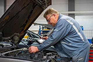 Ein Mechaniker in blauer Arbeitskleidung beugt sich über den geöffneten Motorraum eines Autos und bedient ein Diagnosegerät