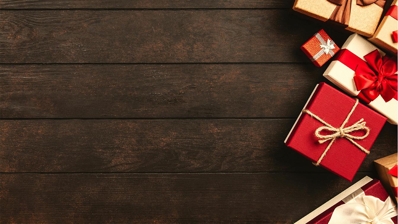 Das Bild zeigt eine festliche Komposition von Weihnachtsgeschenken auf einer rustikalen Oberfläche aus dunkelbraunem Holz. Die Holzbretter verlaufen horizontal. Der Großteil der linken und oberen Fläche bleibt frei, was den Blick auf die Ansammlung von Geschenken am rechten und unteren Rand lenkt. Das auffälligste Element ist ein quadratisches, leuchtend rotes Geschenkpaket in der unteren rechten Mitte, das mit einer einfachen, beigefarbenen Kordelschleife verziert ist. Rechts daneben und darüber sind weitere Geschenke gestapelt: darunter ein weißes Kästchen mit einer großen, roten Satin-Schleife, mehrere naturfarbene oder goldbraune Pakete sowie ein kleines, rotes Päckchen mit einem silbernen Stern. Die Beleuchtung ist gedämpft und erzeugt eine warme, weihnachtliche Stimmung.