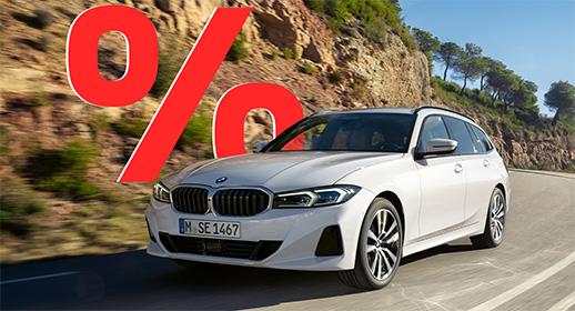 Das Bild zeigt einen weißen BMW 3er-Reihe Touring (Kombi), der auf einer kurvigen Straße fährt. Das Auto ist schräg von vorne zu sehen. Im Vordergrund ist ein großes, rotes Prozentzeichen (%) platziert. Der Hintergrund zeigt eine bergige Landschaft mit Bäumen und Sträuchern. Das Kennzeichen "M SE 1467" ist auf der vorderen Stoßstange zu sehen.