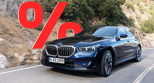  Das Bild zeigt ein weißes, viertüriges SUV der BMW 5er-Reihe Touring, das auf einer Landstraße fährt. Das Auto ist schräg von vorne zu sehen. Im Vordergrund ist ein großes, rotes Prozentzeichen (%) platziert. Der Hintergrund zeigt eine bergige Landschaft mit Windrädern unter einem blauen Himmel.