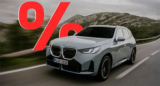  Das Bild zeigt einen hellgrauen, viertürigen BMW X3, der auf einer Landstraße fährt. Das Auto ist schräg von vorne zu sehen. Im Vordergrund ist ein großes, rotes Prozentzeichen (%) platziert. Der Hintergrund zeigt eine bergige Landschaft unter einem bewölkten Himmel. Das Kennzeichen "M XX 3155" ist auf der vorderen Stoßstange zu sehen.          Analyse