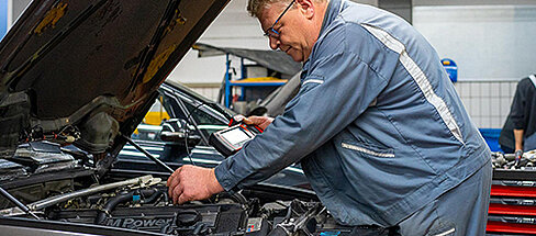 Ein Mechaniker in Arbeitskleidung beugt sich über den offenen Motorraum eines Autos und bedient ein Diagnosegerät. Im oberen linken Bereich des Bildes steht "KARRIERE bei Stadel" und ein Button "Offene Stellen