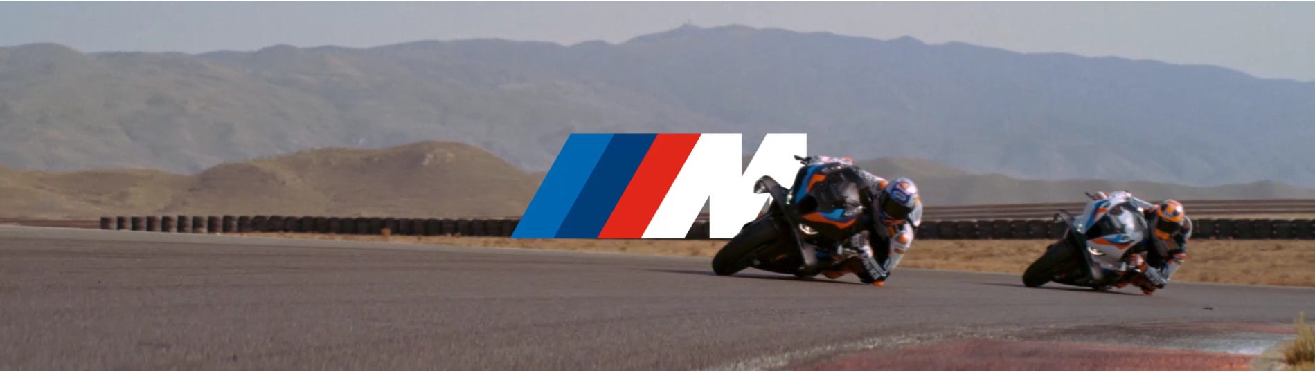 Das Bild zeigt zwei Rennmotorräder auf einer Rennstrecke, die in einer bergigen Wüstenlandschaft liegt. Die Fahrer neigen sich in einer Kurve stark in Richtung der Strecke. Über den Motorrädern schwebt das BMW M Logo in den Farben Blau, Dunkelblau und Rot.