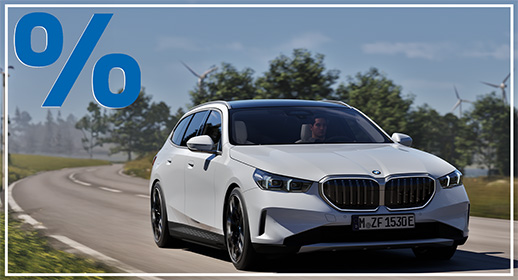 Das Bild zeigt ein weißes, viertüriges SUV der BMW 5er-Reihe Touring, das auf einer Landstraße fährt. Das Auto ist schräg von vorne zu sehen. Im Vordergrund ist ein großes, rotes Prozentzeichen (%) platziert. Der Hintergrund zeigt eine bergige Landschaft mit Windrädern unter einem blauen Himmel.