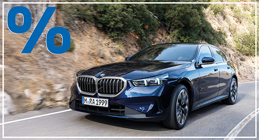  Das Bild zeigt ein weißes, viertüriges SUV der BMW 5er-Reihe Touring, das auf einer Landstraße fährt. Das Auto ist schräg von vorne zu sehen. Im Vordergrund ist ein großes, rotes Prozentzeichen (%) platziert. Der Hintergrund zeigt eine bergige Landschaft mit Windrädern unter einem blauen Himmel.