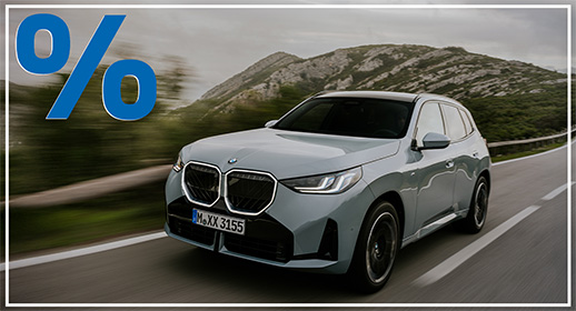Das Bild zeigt einen hellgrauen, viertürigen BMW X3, der auf einer Landstraße fährt. Das Auto ist schräg von vorne zu sehen. Im Vordergrund ist ein großes, rotes Prozentzeichen (%) platziert. Der Hintergrund zeigt eine bergige Landschaft unter einem bewölkten Himmel. Das Kennzeichen "M XX 3155" ist auf der vorderen Stoßstange zu sehen.          Analyse