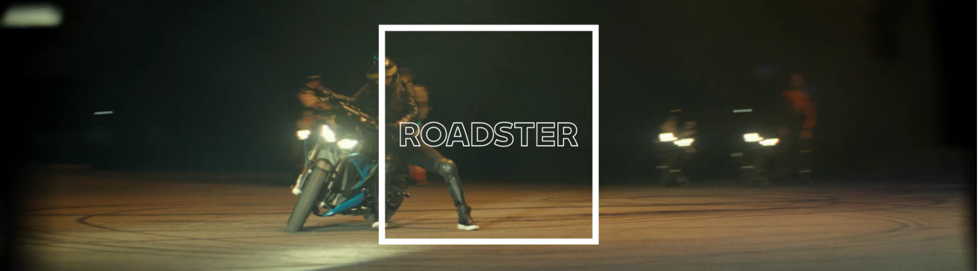 Ein Bild in einem breiten Panorama-Format, das eine nächtliche Szene zeigt. Im Vordergrund steht ein Motorradfahrer in Lederkleidung neben seinem blauen Roadster-Motorrad. Der Scheinwerfer des Motorrads ist an und leuchtet nach vorne. Der Fahrer stützt sich mit einem Fuß auf den Boden. Im Hintergrund sind undeutlich weitere Motorräder mit brennenden Scheinwerfern zu erkennen. Das gesamte Bild ist von einem schmalen, weißen Rahmen umgeben, in dem das Wort „ROADSTER“ in Großbuchstaben steht.